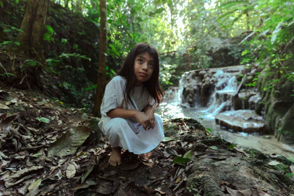 A young girl in traditional clothing explores a peaceful forest by a flowing waterfall.